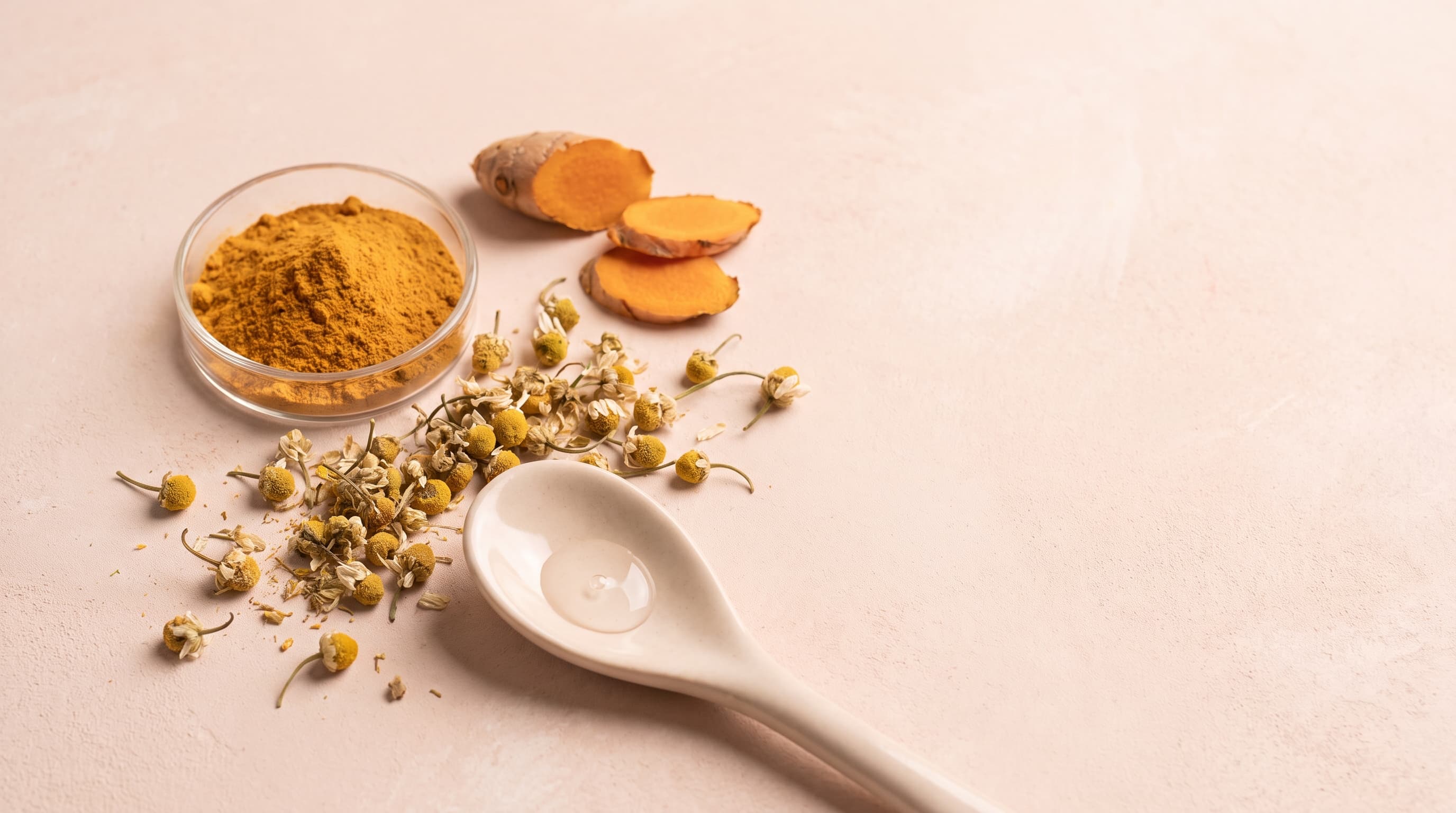 Ground turmeric, fresh turmeric root and chamomile flowers on a soft pink surface